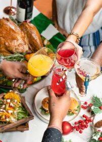 The painting depicts a festive meal, likely a Christmas dinner or similar feast, with a roasted bird (probably turkey or chicken), various side dishes, and people clinking glasses to toast.