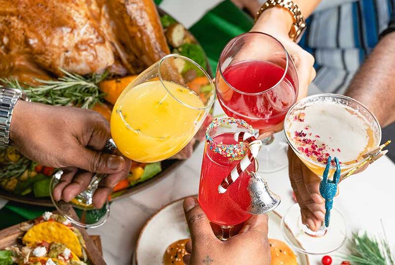 The painting depicts a festive meal, likely a Christmas dinner or similar feast, with a roasted bird (probably turkey or chicken), various side dishes, and people clinking glasses to toast.